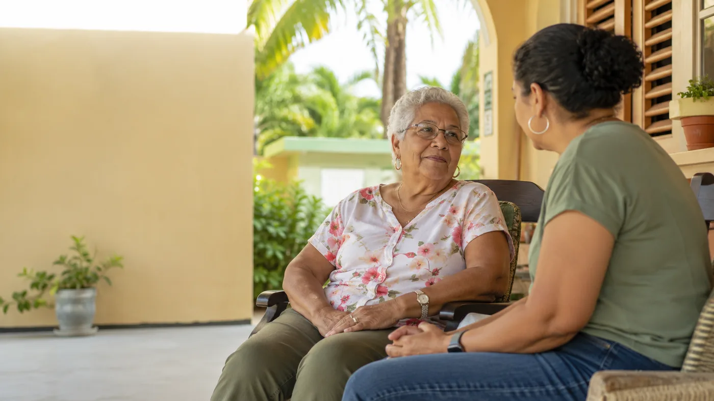 Un señora mayor y un mucha muhé sinta huntu na un veranda cu solo, papiando